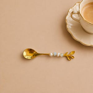 May include: A gold-toned metal spoon with a handle featuring a branch-like design adorned with four small, irregular white and pale blue beads. The spoon is positioned horizontally next to a matching gold-rimmed ceramic cup filled with coffee.