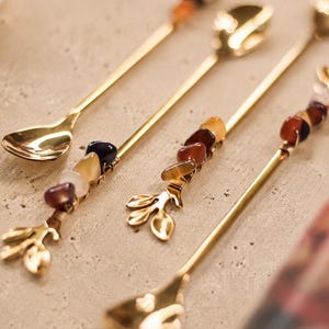 Puede incluir: Un juego de cucharas de metal en tono dorado con mangos largos y delgados. Cada mango está adornado con un grupo de cuentas de piedras preciosas pulidas y multicolores sujetas con alambre fino. Los extremos tienen forma de pequeñas ramas de tres hojas.