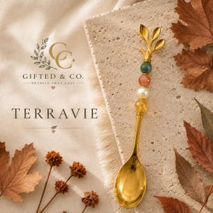 Puede incluir: Una cuchara de metal dorado con un mango decorativo rematado con un diseño de tres hojas y cuatro cuentas esféricas en verde, naranja, blanco y amarillo. La cuchara está colocada verticalmente sobre una piedra texturizada junto a hojas de otoño.