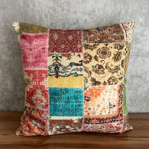 May include: Square patchwork pillow with a variety of patterned fabric squares in shades of red, pink, teal, yellow, and brown. The pillow is placed on a wooden surface against a gray background.