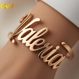 以下が含まれることがあります： 筆記体で「Valeria」と書かれたゴールドカラーのカフブレスレット。オープンエンドのブレスレットは手首に着用されています。盛り上がった文字は光を反射する研磨仕上げです。全体的なデザインはエレガントでパーソナライズされています。