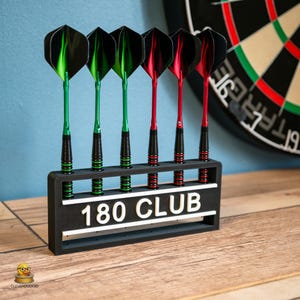 Op de afbeelding: Een zwarte rechthoekige dartstandaard met de witte tekst 180 CLUB op de voorzijde. De standaard bevat zes darts, drie met groene flights en shafts, en drie met rode flights en shafts, allemaal voorzien van zwarte, getextureerde grepen.