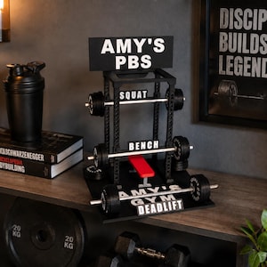 Puede incluir: Un expositor en miniatura negro de jaula de potencia con el texto AMY'S PBS en la parte superior, etiquetas SQUAT, BENCH y DEADLIFT en la estructura, un banco rojo y barras plateadas con discos negros. La base tiene la inscripción AMY'S GYM.