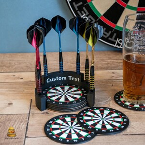 May include: A black circular dartboard-themed coaster holder with the text Custom Text, containing a stack of dartboard coasters. Six metal darts with colourful flights in red, blue, and yellow are inserted into the holder's top rim.