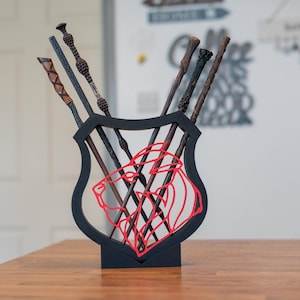 Könnte beinhalten: Ein schwarzer Zauberstabhalter in Schildform mit einem roten Löwenemblem. Mehrere Zauberstäbe in verschiedenen Farben und Designs werden darin präsentiert, die auf einem Holztisch liegen. Die Zauberstäbe weisen komplizierte Details und Texturen auf.