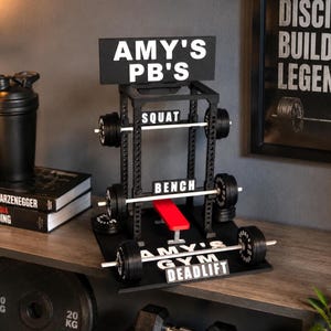 May include: A black miniature gym rack display featuring white text that reads AMY'S PB'S, SQUAT, BENCH, AMY'S GYM, and DEADLIFT. The structure includes three silver barbell rods with black weight plates and a small red bench centered in the rack.