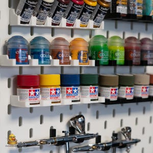 May include: A collection of model paints in various colors, including red, yellow, blue, and green, displayed on a white pegboard. The image also shows airbrushes, and paint tubes. The paints are organized on shelves, with the brand names visible.