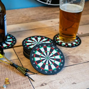 May include: A set of circular drink coasters featuring a dartboard design with black, white, red, and green segments and numbered markings. The coasters are arranged in a stack and individually, with two yellow and black darts placed nearby.