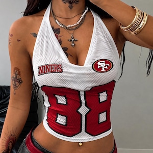 May include: A white mesh halter top featuring the red text NINERS, a red and gold San Francisco 49ers logo, and the large red number 88 on the front. The garment has a deep neckline and is constructed from a perforated athletic fabric.