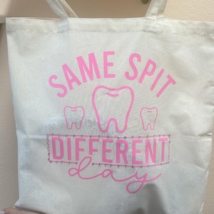 May include: Off-white canvas tote bag with pink text and tooth graphics. The text reads "SAME SPIT DIFFERENT DAY". The bag has two handles and is held up by a person's hand.