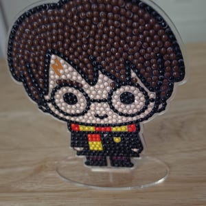 May include: A decorative acrylic standee featuring a pixelated character design made of small, round, multi-colored beads. The figure has dark brown hair, round glasses, a lightning bolt mark on the forehead, and a black robe with a red and yellow scarf.