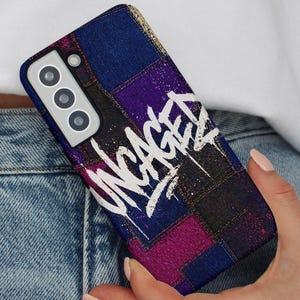 May include: A phone case with a patchwork design in shades of blue, purple, and pink. The case features the word "UNCAGED" in white graffiti-style lettering. The phone case has a camera module with three lenses.
