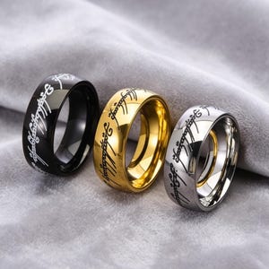 May include: Three rings in black, gold, and silver, each with engraved script. The rings are displayed on a gray surface. The gold ring has a shiny finish, while the black and silver rings have a polished look.