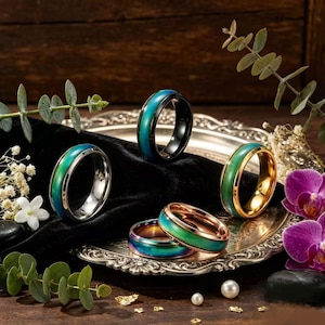 Op de afbeelding: Een collectie van vijf stemmingsringen met van kleur veranderende banden in tinten groen en blauw. De ringen hebben metalen afwerkingen in zilver, goud, roségoud en iriserende tinten, uitgestald op een zilveren dienblad met zwarte fluwelen stof.