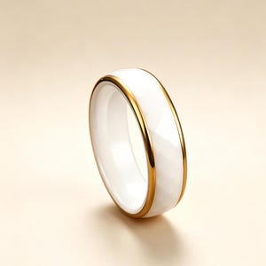 May include: A white ceramic ring with a faceted surface and a gold-colored band. The ring has a simple, elegant design. The ring is a jewelry item.