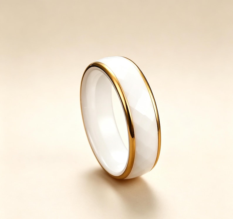 May include: A white ceramic ring with a faceted surface and a gold-colored band. The ring has a simple, elegant design. The ring is a jewelry item.