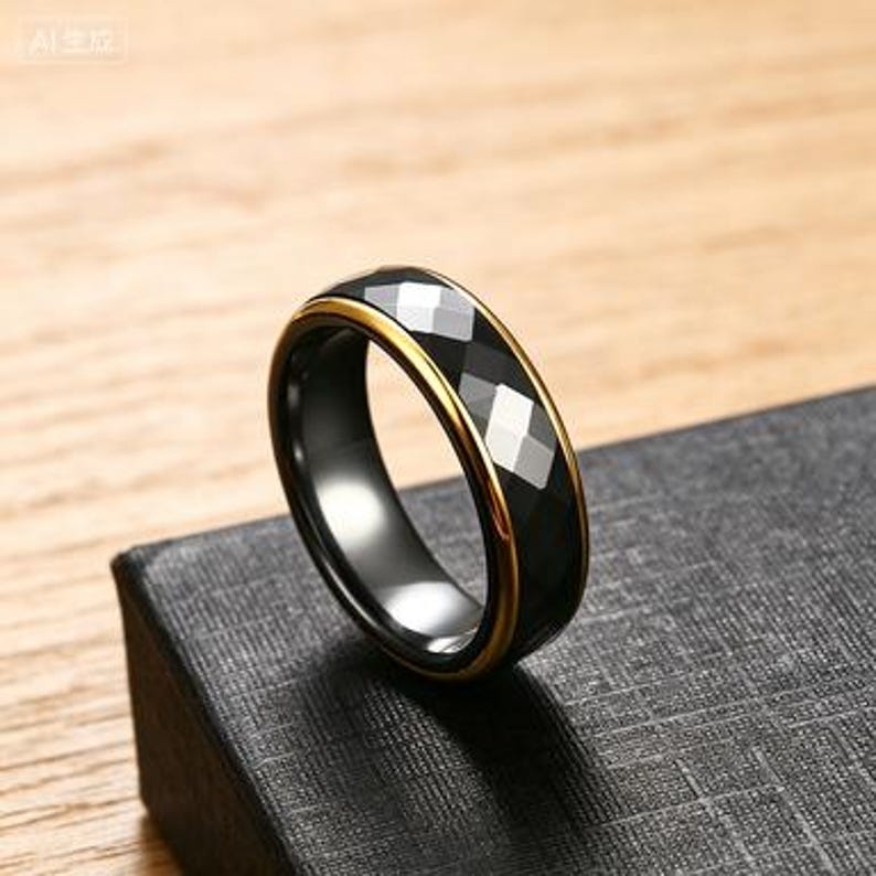 May include: A black ring with a faceted, geometric design and a gold-colored band. The ring is set on a black box, with a wooden surface in the background.