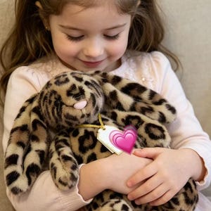 May include: A plush leopard-print bunny with long floppy ears and a pink nose. The stuffed animal has a tag with a pink heart attached. The bunny is being held by a person wearing a pink dress.