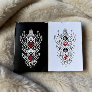 Puede incluir: Dos barajas de cartas, una negra y otra blanca, cada una con un diseño simétrico con motivos de corazones, picas, tréboles y diamantes en rojo y blanco. Las cartas están sobre una superficie suave y clara.