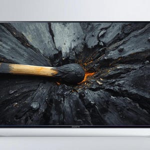 May include: A flat-screen television with a black frame and silver legs. The screen displays a close-up of a burnt match on a dark, textured surface, with orange embers visible. The image has a dramatic, high-contrast aesthetic.