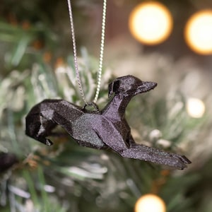 May include: A black, geometric, low-poly style whippet dog ornament hanging from a thin silver string. The figure features sharp, angular facets and is suspended against a blurred background of green pine needles and warm, glowing bokeh fairy lights.