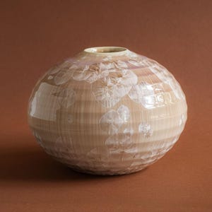 Könnte beinhalten: Eine kugelförmige Keramikvase mit einer hellbeigen, gerippten Oberfläche und weißen kristallinen Glasurmustern. Die Vase hat eine kleine, kreisförmige Öffnung an der Oberseite und einen abgerundeten Körper, der sich zur Basis hin verjüngt.