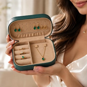 Op de afbeelding: Een tealkleurige reisetui voor sieraden, open om verschillende stukken te tonen. De etui bevat gouden oorbellen, ringen en een ketting, allemaal met groene edelstenen. De binnenkant is lichtbeige.