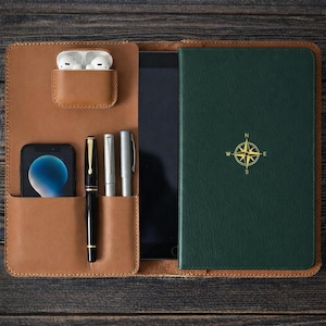 May include: A brown leather folio with pockets for a smartphone, pens, and wireless earbuds. A green notebook with a compass rose design is visible. The folio holds a tablet. The items are arranged on a dark wood surface.