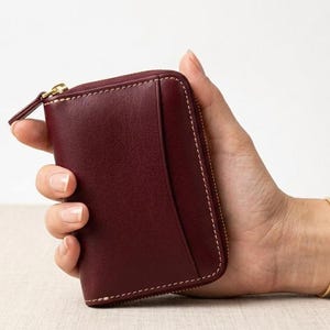 May include: A burgundy leather wallet with a gold zipper, held in a hand. The wallet has white stitching and a small pocket on the side. The background is a neutral color.