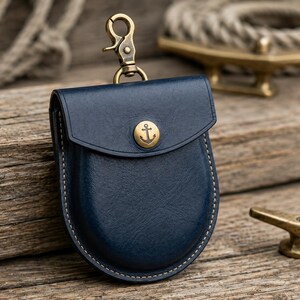 May include: A navy blue leather pouch with a gold-colored anchor button and a matching clasp. The pouch has white stitching around the edges and is designed to hold small items. Nautical theme.