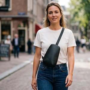 May include: A woman wearing a white t-shirt and blue jeans, with a black crossbody bag. The bag is worn across the body. The background shows a European street scene with brick buildings and people walking.