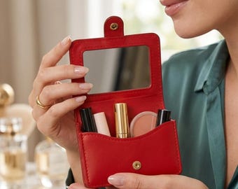 May include: A red leatherette makeup case with a mirror and various cosmetic products. The case is held open, revealing a compact mirror, lipstick, and other beauty items. The case is rectangular with a snap closure.