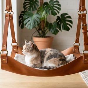 Pode incluir: Uma rede para gatos em couro castanho, suspensa por correias com ferragens de latão. Um gato cinza e branco descansa dentro da rede. Uma planta em vaso está ao fundo.