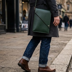 May include: A dark green leather messenger bag with a diagonal flap closure and a shoulder strap. The bag is carried by a person wearing a grey coat, blue jeans, and brown leather shoes. The bag appears to be approximately 30 cm tall and 25 cm wide.