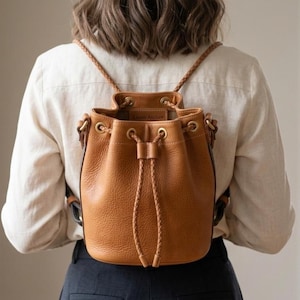 以下が含まれることがあります： 編み込みのショルダーストラップと巾着開閉式のタンレザーバケットバックパック。 バッグにはゴールドカラーの金具と開口部内側に小さなタグが付いています。 バッグは背中に着用されています。