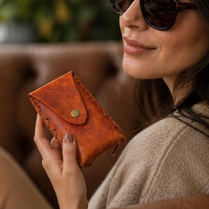 May include: A handcrafted, orange leather glasses case with a flap closure and a brass button. The case has decorative stitching along the edges. The case is designed to protect eyeglasses and sunglasses.