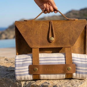 May include: A brown leather picnic bag with a flap closure and handle. The bag holds a striped towel secured by leather straps. The text "Lines Atölyesi" is visible on the strap. The bag is held up against a scenic background.