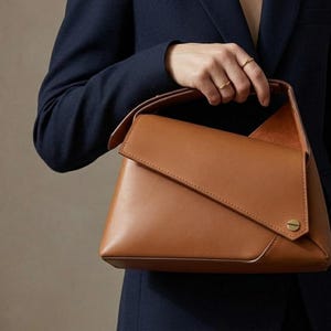 Puede incluir: Un bolso de mano de cuero color marrón con un diseño geométrico. El bolso tiene un cierre de solapa y un pequeño acento circular dorado. El asa es un trozo de cuero doblado. El bolso lo lleva una persona con una blazer azul marino.