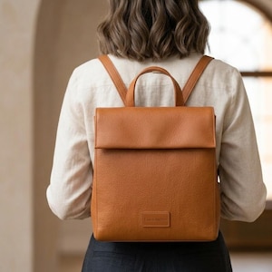 Puede incluir: Una mochila de cuero marrón con cierre de solapa y asa superior. La mochila tiene correas ajustables y una pequeña etiqueta rectangular en la parte delantera. La bolsa se lleva en la espalda de una persona que lleva una camisa clara.