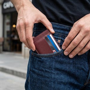 May include: A burgundy leather wallet with visible credit cards is being placed into the front pocket of blue denim jeans. The wallet has a minimalist design with visible stitching. The person is wearing a black t-shirt.