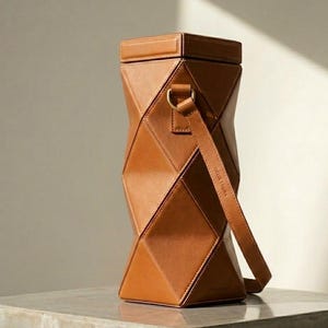 Puede incluir: Un portabotellas de vino geométrico de cuero marrón con una correa para el hombro. El portabotellas tiene un diseño angular único con una parte superior rectangular y un cuerpo cilíndrico. La correa está sujeta con un anillo dorado. El texto "HELIX TRUNK" está impreso en la correa.