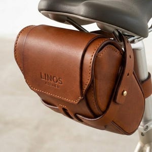 Op de afbeelding: Bruine leren fietstas bevestigd aan de zadelpen. De tas is voorzien van de tekst "LINOS ATELIER". De tas is gemaakt van rijk, getextureerd leer en heeft een klassiek ontwerp.
