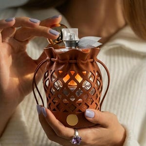May include: A brown leather perfume bottle holder with a diamond-patterned cutout design. The perfume bottle is visible through the cutouts. The holder has a drawstring closure and a gold-colored accent. A light blue ribbon is tied around the bottle's neck.