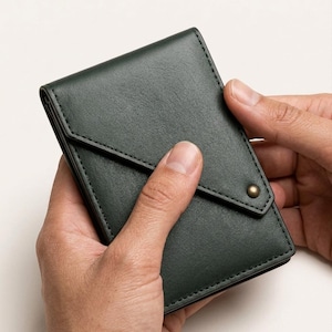 May include: Dark green leather wallet with a flap closure and a brass-colored snap. The wallet is held open, showcasing its interior. The stitching is a contrasting black color.