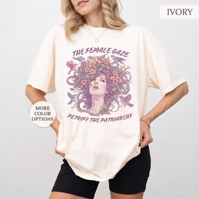 Puede incluir: Camiseta de color marfil con un gr&aacute;fico de un rostro femenino con cabello floral, p&aacute;jaros y el texto "THE FEMALE GAZE PETRIFY THE PATRIARCHY". La camiseta tiene mangas cortas y un corte holgado. "MORE COLOR OPTIONS" est&aacute; impreso en el lado izquierdo.