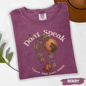 May include: Berry-colored t-shirt with the words "Don't Speak" and a botanical illustration of a pomegranate and rose. The shirt is a Comfort Colors brand, with a crew neck and short sleeves.