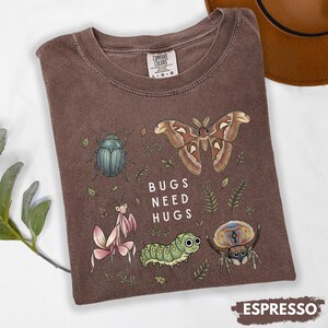 May include: A brown short-sleeve t-shirt featuring a graphic print of a beetle, a moth, a praying mantis, a caterpillar, and a spider. The text "BUGS NEED HUGS" is printed in white in the center. The shirt has a crew neckline and a label.