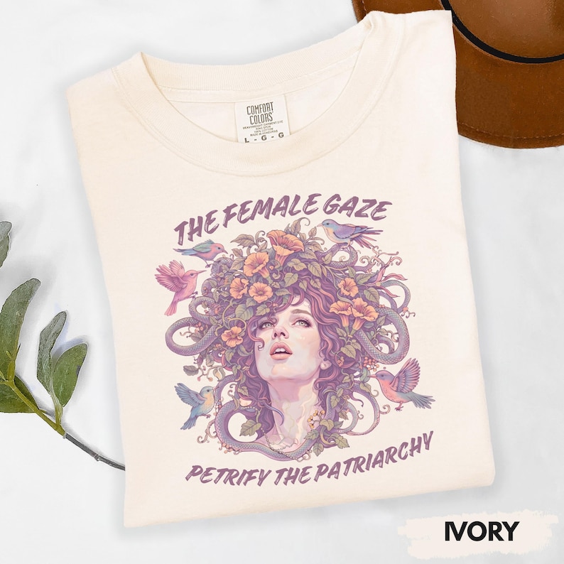 Puede incluir: Camiseta color marfil con el texto "THE FEMALE GAZE" y "PETRIFY THE PATRIARCHY". El dise&ntilde;o presenta un rostro femenino con elementos florales y serpientes, rodeado de p&aacute;jaros. Marca Comfort Colors, talla L-G.