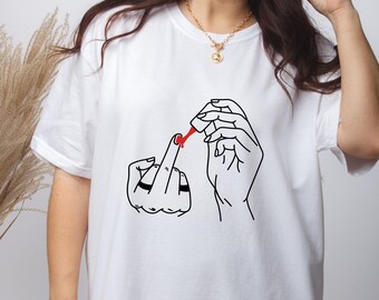 May include: White t-shirt featuring a black line drawing of hands applying red nail polish to a middle finger. The design is centered on the shirt. The shirt has short sleeves and a crew neck.