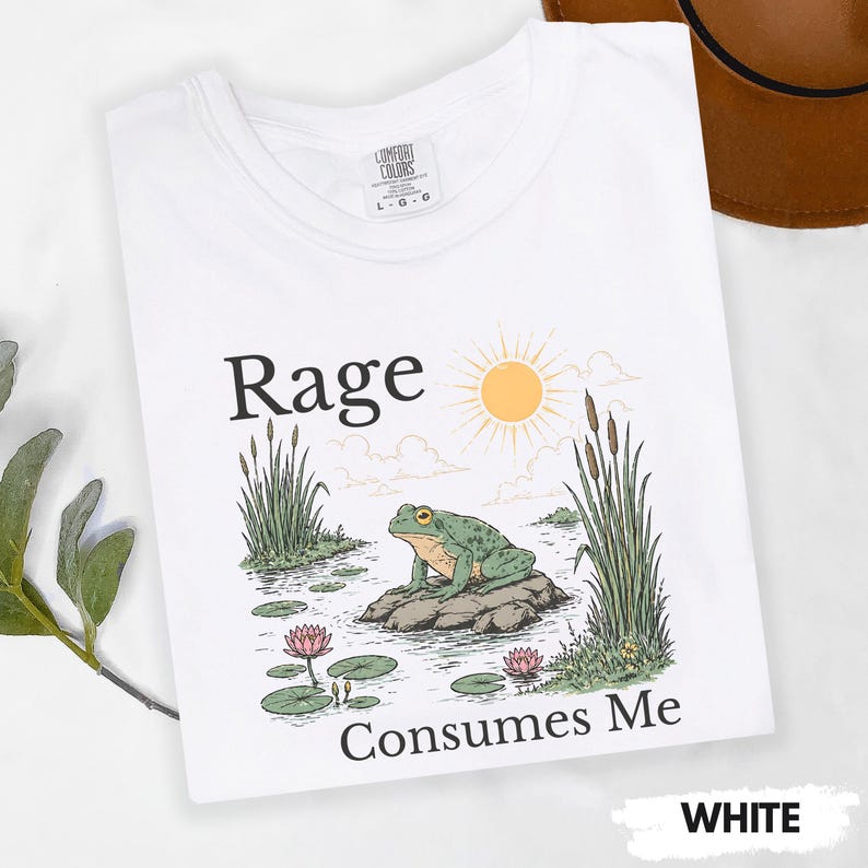 Puede incluir: Camiseta blanca con el texto "Rage Consumes Me" y un gr&aacute;fico de una rana verde sobre una roca en un estanque con nen&uacute;fares y juncos. El sol est&aacute; en el fondo. La camiseta est&aacute; etiquetada como "Comfort Colors".